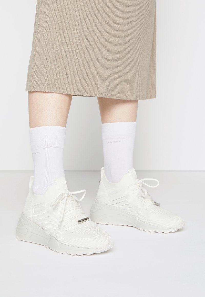 White athletic sneakers with a textured mesh upper, lace-up design, and raised rubber sole, paired with white crew socks and a beige ribbed skirt.