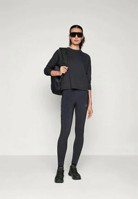 Crna dukserica, tamne tajice visoko strukirane i crne tenisice. Dodaci uključuju crni ruksak i sunčane naočale. Glatki, prilagođeni materijali.