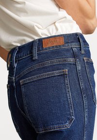 Mörkblå denimjeans med slim fit, utrustade med en brun läderetikett vid midjan och dubbla bakfickor med kontrasterande sömmar.