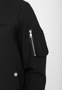 Manche de veste noire avec une petite poche zippée et une poche à rabat adjacente sur le haut du bras sur un fond uni.