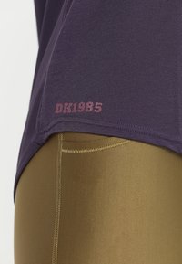 Tumman violetin puuvillapaita, jossa on pieni "DK1985" -painatus maroonilla helman kohdalla, yhdistettynä kiiltäviin kultaisiin legginsseihin, joilla on sileä pinta.