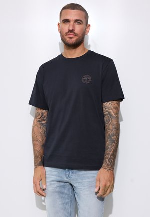 Mand med kort hår og skæg iført sort t-shirt og lyseblå jeans, der viser tatoverede full sleeves på begge arme.