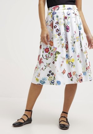 Frau trägt weißen Midirock mit buntem Blumenmuster und schwarze flache Schuhe mit Knöchelriemen, steht vor schlichtem Hintergrund.