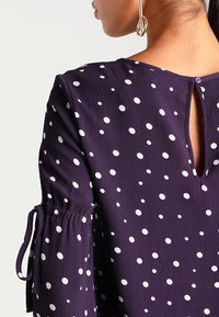 Blusa viola con pois bianchi, maniche elasticizzate con lacci e apertura a chiave sul retro con dettaglio di bottone. Tessuto texturizzato.