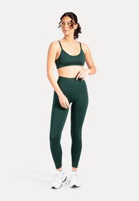 Donkergroene sportbeha en leggings met hoge taille gemaakt van gladde stof. Model draagt witte sneakers en staat tegen een lichte achtergrond.