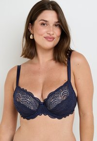 Soutien-gorge en lace bleu marine avec armatures, présentant un bord festonné, un motif floral détaillé et des bretelles réglables. Petit accent en nœud au centre.