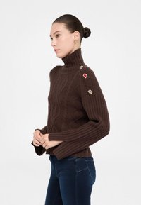 Maglione marrone lavorato a maglia con collo alto, maniche a coste e bottoni decorativi sulla spalla. Motivo a treccia testurizzato su tutta la superficie.