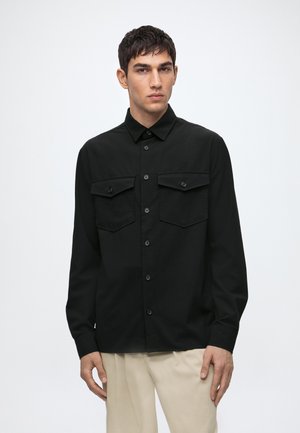 Homme portant une chemise noire boutonnée avec deux poches poitrine et un pantalon beige, debout devant un fond clair uni.