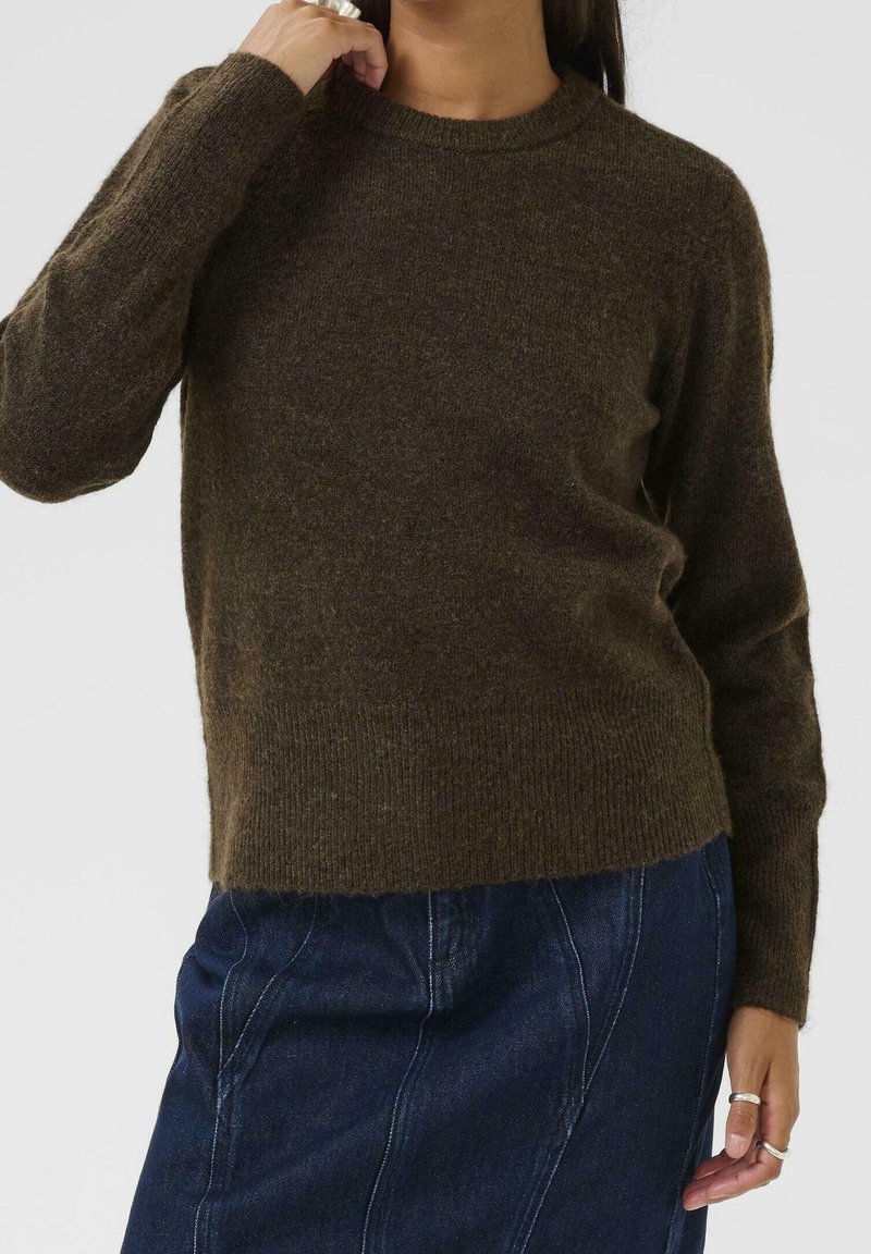 Personne portant un pull à manches longues marron foncé et une jupe en denim bleu, avec la main touchant le cou et des bagues visibles aux doigts.