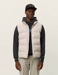 Ανοιχτό γκρίζο γιλέκο με φερμουάρ, διαθέτει κουκούλα και πλαϊνές τσέπες, φορεμένο πάνω από ένα μαύρο φούτερ. Το μοντέλο φοράει ένα μαύρο καπέλο.