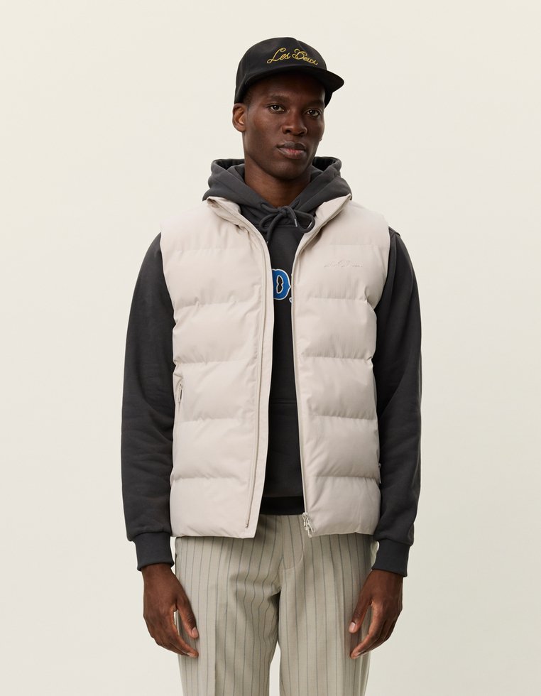 Ανοιχτό γκρίζο γιλέκο με φερμουάρ, διαθέτει κουκούλα και πλαϊνές τσέπες, φορεμένο πάνω από ένα μαύρο φούτερ. Το μοντέλο φοράει ένα μαύρο καπέλο.