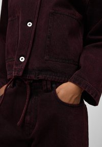 Giacca di denim bordeaux con un design cropped, due tasche frontali e chiusura con bottoni a pressione; pantaloni abbinati con coulisse visibili in vita.