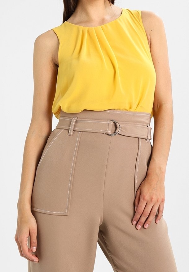Femme portant un chemisier jaune sans manches rentré dans un pantalon beige taille haute avec une large ceinture et une grande poche avant.
