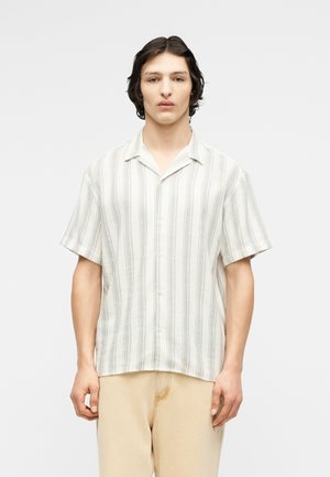 Νέος άνδρας όρθιος, φορούντας ένα κοντομάνικο λευκό πουκάμισο με κατακόρυφες γκρι ρίγες και μπεζ παντελόνι, σε λευκό φόντο.