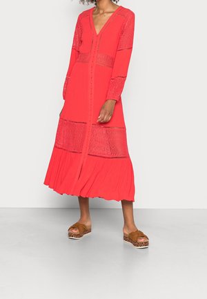 Femme portant une robe midi rouge avec des panneaux en dentelle, des manches longues, des boutons recouverts de tissu sur le devant, et des sandales compensées marron avec un détail nœud.