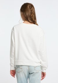 Felpa bianca con una vestibilità rilassata, maniche lunghe e polsini a coste. Texture morbida e orlo elasticizzato. Indossata con jeans azzurri.