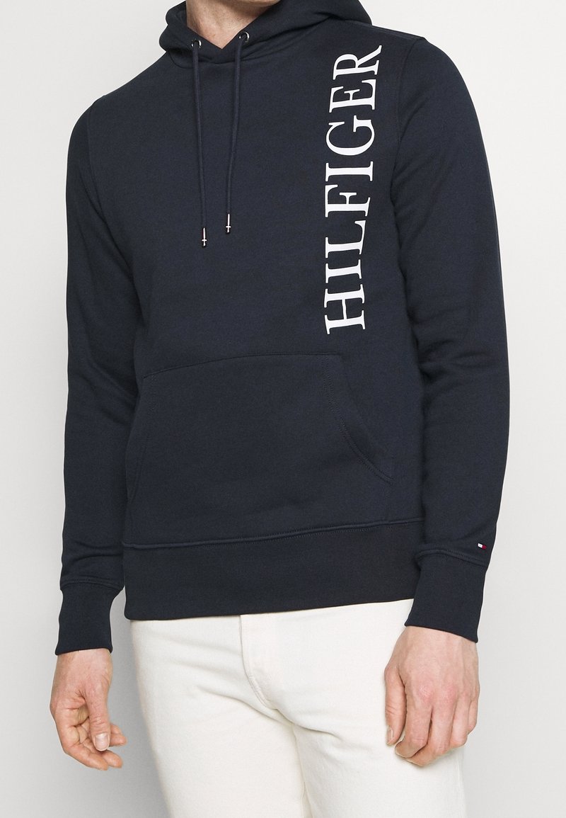 Homme portant un sweat à capuche noir avec le texte vertical blanc « HILFIGER » sur la poitrine et un pantalon blanc, debout devant un fond uni.