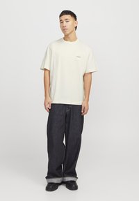 Lys beige kortermet T-skjorte med avslappet passform, med en liten logo på venstre bryst. Kombinert med mørkeblå vide jeans og svarte sko.