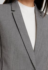 Schwarz-weißer kariierter Blazer mit Reverskragen. Getragen über einem weißen Oberteil mit glatter Textur und taillierter Passform.