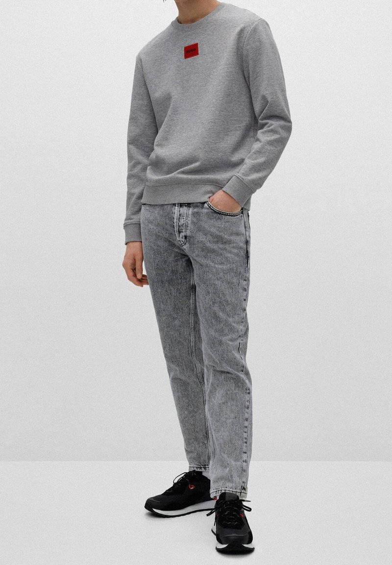 Personne portant un sweat-shirt gris avec un logo rouge, un jean gris clair délavé à l'acide, et des baskets noires, debout sur fond blanc.
