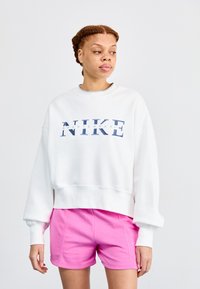 Bijela skraćena dukserica s grafikama "NIKE SPORTSWEAR" u plavoj boji, u kombinaciji s ružičastim sportskim šortsevima. Glatka tkanina, širok kroj, okrugli izrez.