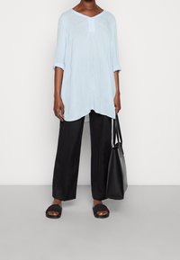 Person in einem hellblauen Langarm-Tunikakleid, mit schwarzen weiten Hosen, schwarzen Sandalen und einer großen schwarzen Tragetasche vor einem schlichten Hintergrund.