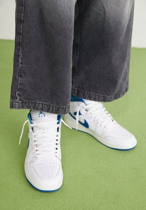 White high-top sneakers with blue accents, perforated toe box, and white laces. Worn with loose gray denim pants on a green surface.