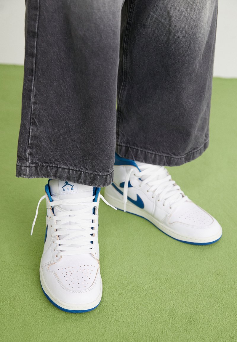 Białe wysokie sneakersy z niebieskimi akcentami, perforowanym czubkiem i białymi sznurowadłami. Noszone z luźnymi szaro-denimowymi spodniami na zielonej powierzchni.