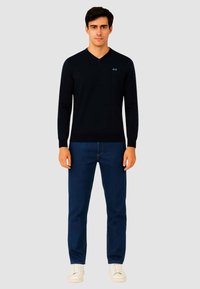 Maglione nero a scollo a V realizzato in morbido tessuto jersey, abbinato a jeans in denim blu scuro e sneakers bianche. Presenta un discreto dettaglio del logo sul petto.