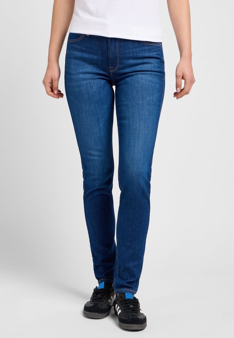 Personne portant un jean skinny bleu, des baskets noires à rayures blanches et une chemise blanche, debout sur un fond uni gris clair.