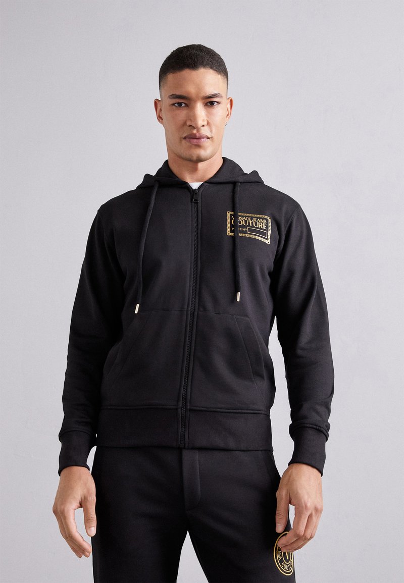 Zwarte zip-up hoodie van zachte stof, met een voorlogo patch, trekkoorden met metalen punten en een kangoeroezak.