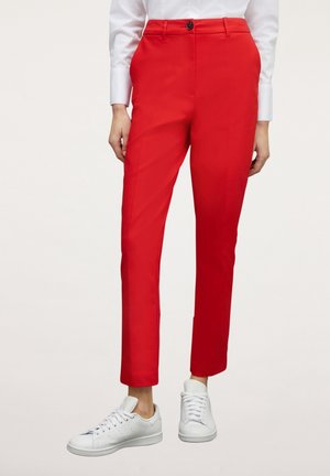 Slim-fit rode broek gedragen met witte sneakers en een wit shirt met lange mouwen, getoond van midden torso tot voeten.