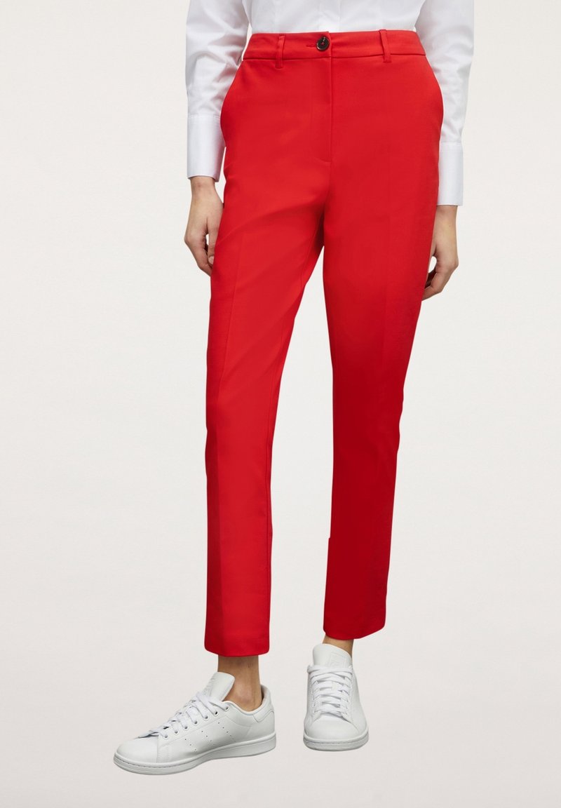 Pantalon rouge slim porté avec des baskets blanches et une chemise blanche à manches longues, montré de la moitié du torse jusqu’aux pieds.