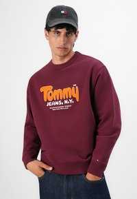 Joven con sudadera burdeos con texto naranja "Tommy Jeans, N.Y." y una gorra oscura, de pie con las manos en los bolsillos de los jeans.