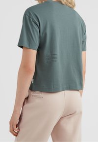 Puuvillainen t-paita hillityssä sinivihreässä sävyssä, lyhyt malli, takana painettu tekstidetalji. Yhdistetty vaaleisiin beigen värisiin housuihin. Sivulla näkyvä merkintä.