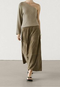 Haut en maille beige à une épaule associé à une longue jupe en daim olive, avec un design élégant et des sandales plates noires. Texture lisse et forme fluide.