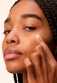 Se está aplicando una base beige en la mejilla de una persona con piel de tono marrón medio. El cabello de la persona está en trenzas largas y bien cuidadas.