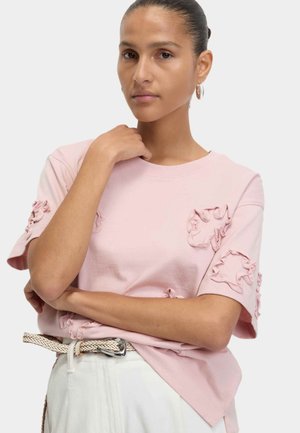 Light pink cotton t-shirt featuring ruffled accents on the sleeves and chest, with a round neckline and relaxed fit. Paired with a woven belt.