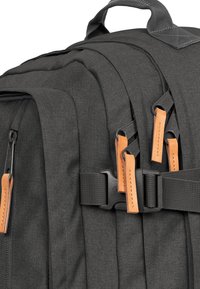 Mochila de tela negra con múltiples compartimentos, con tiradores de cremallera de cuero naranja y herrajes negros. Material texturizado y correas duraderas.