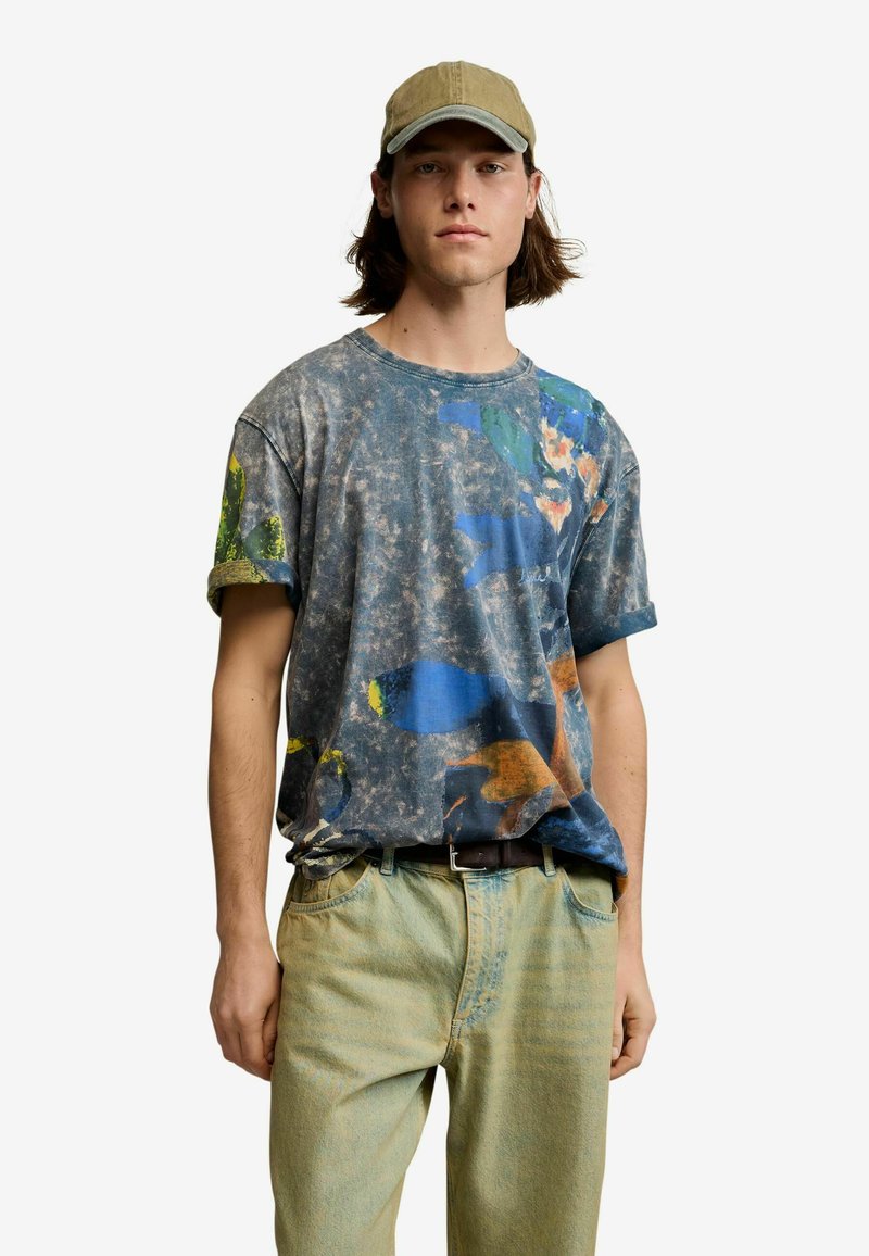 Young man with shoulder-length hair wearing a beige cap, a faded t-shirt with blue and orange abstract patterns, and light green pants.