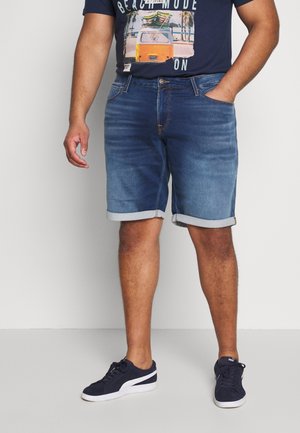 Homme portant un short en jean bleu foncé retroussé, un t-shirt graphique bleu marine et des baskets noires à semelles blanches, debout contre un mur uni.