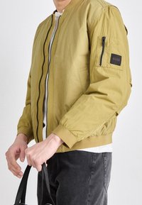 Bomber bunda olivově zelená, s předním zipem, žebrovanými manžetami a lemem a černou látkovou nášivkou na levé horní paži.