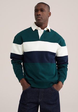 Homme portant un maillot de rugby à manches longues avec des rayures horizontales vertes, blanches et bleu marine, debout les mains dans les poches devant un fond uni.