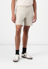 Pantaloni scurți bej cu dungi, cu talie elastică și șnur. Combinati cu șosete negre și adidași gri-albi cu accente negre.