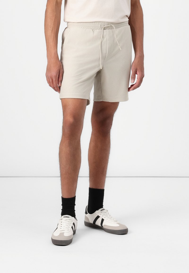 Pantaloni scurți bej cu dungi, cu talie elastică și șnur. Combinati cu șosete negre și adidași gri-albi cu accente negre.