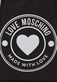 Svart tygväska med en framträdande vit cirkulär logotyp som inkluderar texten "LOVE MOSCHINO" och en hjärtsymbol, med "MADE WITH LOVE" under.