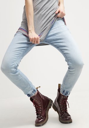 Vaqueros ajustados de mezclilla en azul claro con puños elásticos, combinados con botas de cuero burdeos con cordones que tienen pestañas amarillas y suelas negras.
