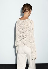 Helles Creme, locker gestrickter Pullover mit langen Ärmeln, strukturiertem Muster und einem gewellten Saum. Über weißen Hosen getragen, von hinten betrachtet.