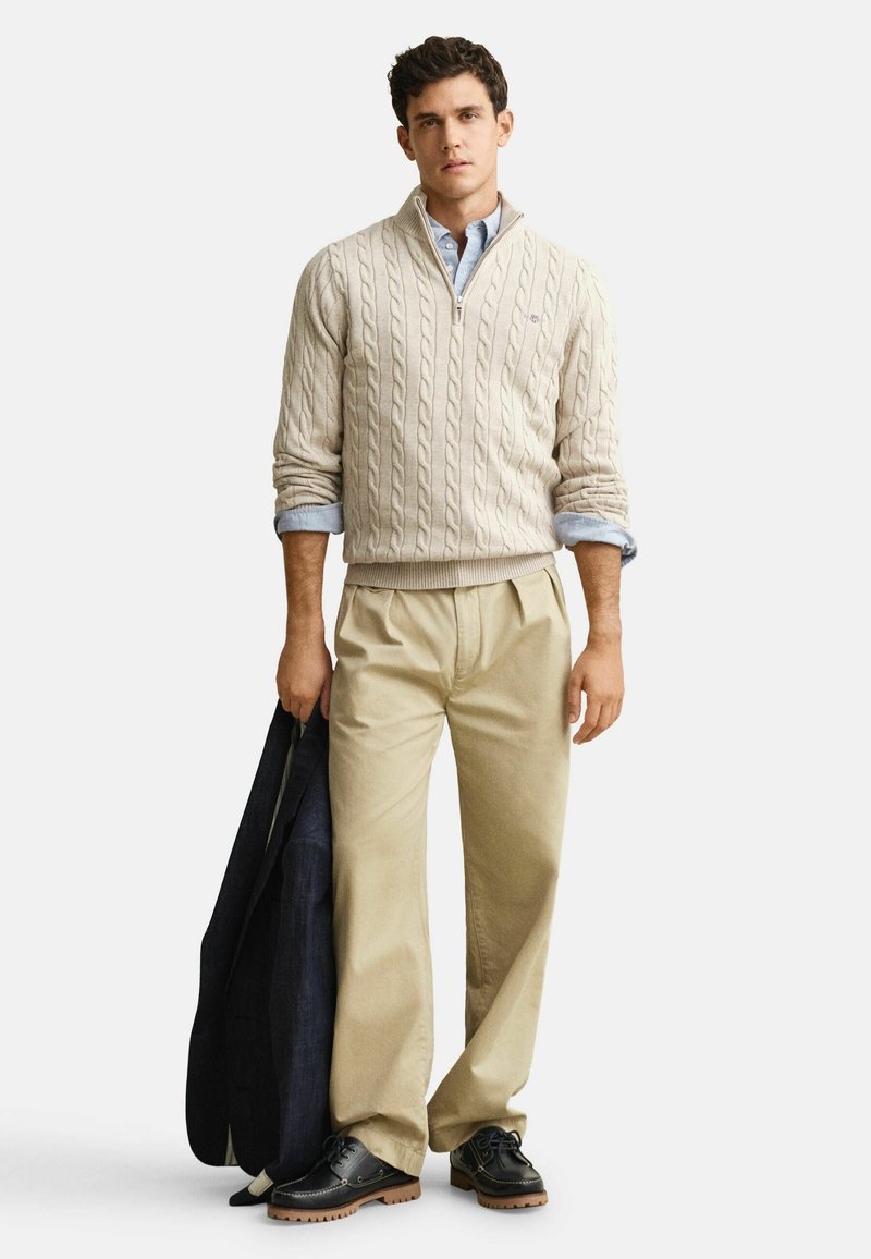 Homme portant un pull beige à col zippé en maille torsadée, chemise bleu clair, pantalon beige plissé, tenant une veste bleu marine, chaussures noires à semelles marron, debout.