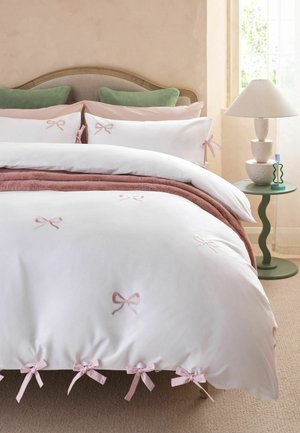 Bett mit weißer und rosa Schleifen geschmückter Bettdecke und Kissen, grünen Kissen, einem grünen Beistelltisch mit Lampe und einem Fenster mit natürlichem Licht.